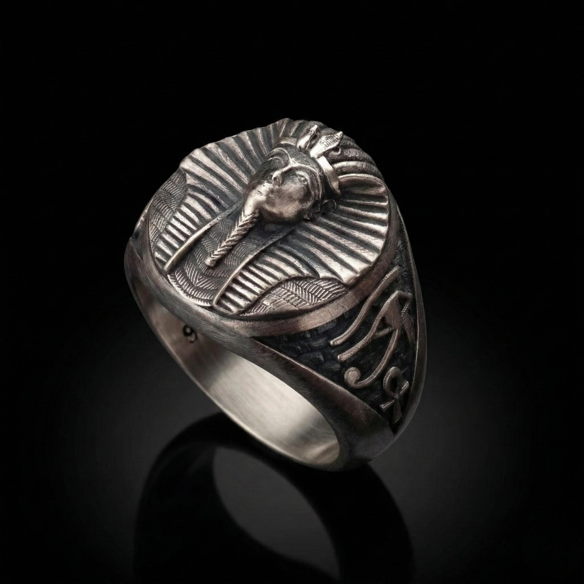 A silver ring with an embossed design featuring a stylized human head or face, possibly a pharaoh or deity, against a black background.