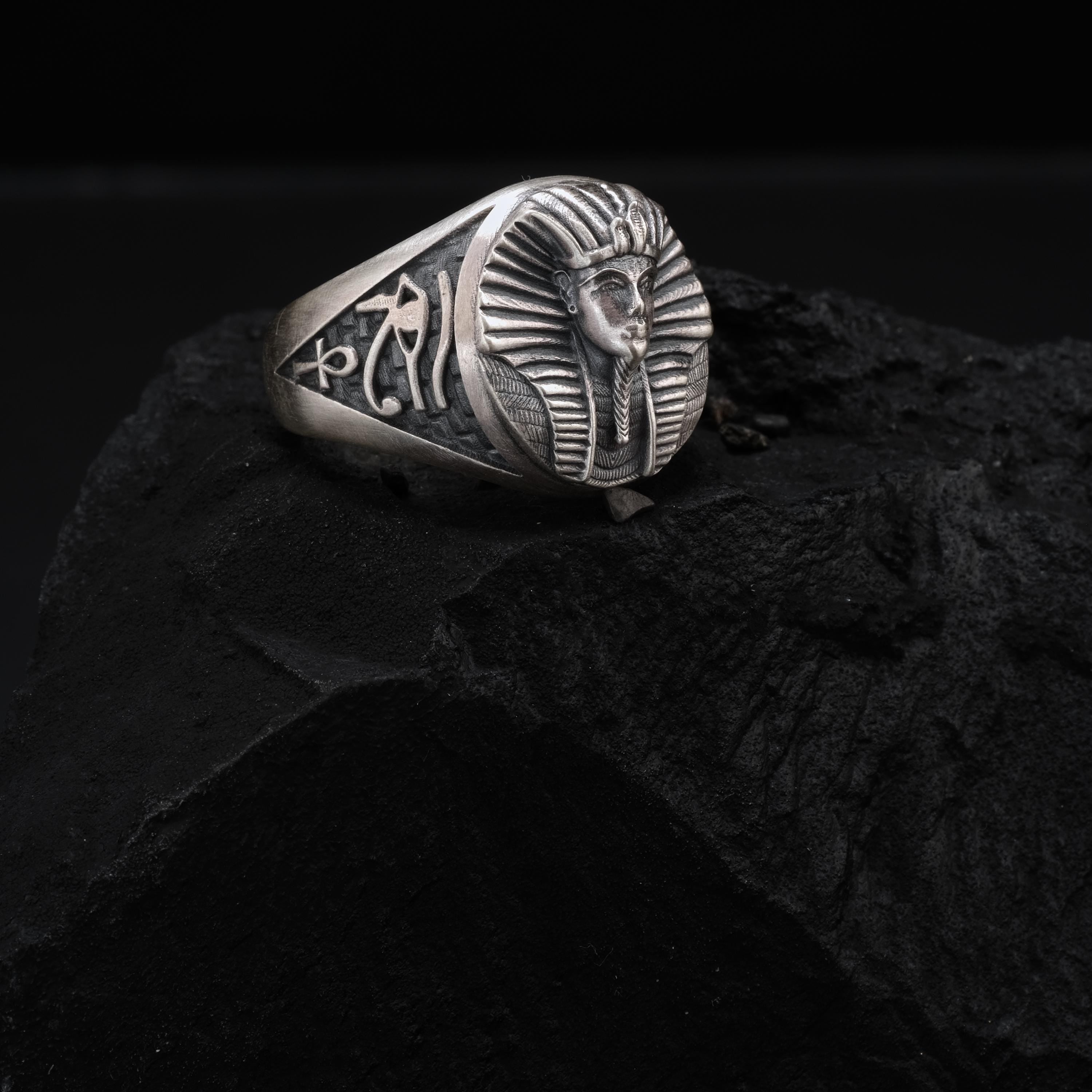 A silver ring with an embossed image of a pharaoh's head, possibly representing the Egyptian god Ankh, is displayed on a dark surface.