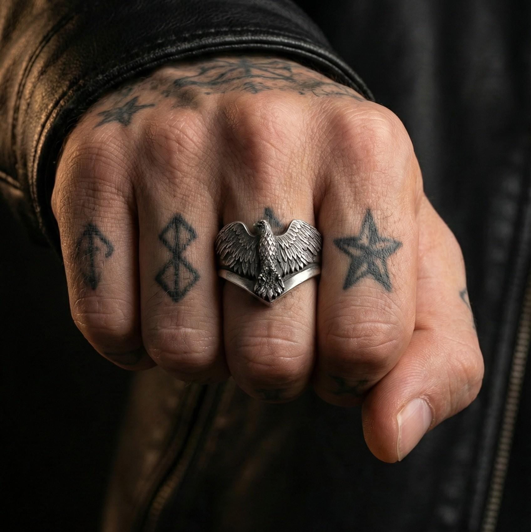 A close-up of a person's hand with multiple tattoos on the fingers, wearing a ring with a bird design.