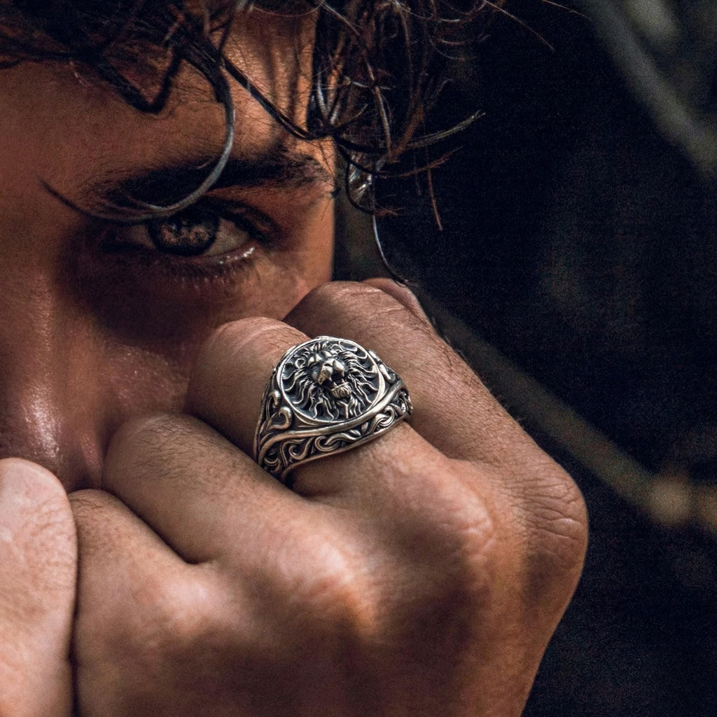 A close-up of a person's face, with their eyes closed and a hand covering their face, wearing a silver ring with a lion design.