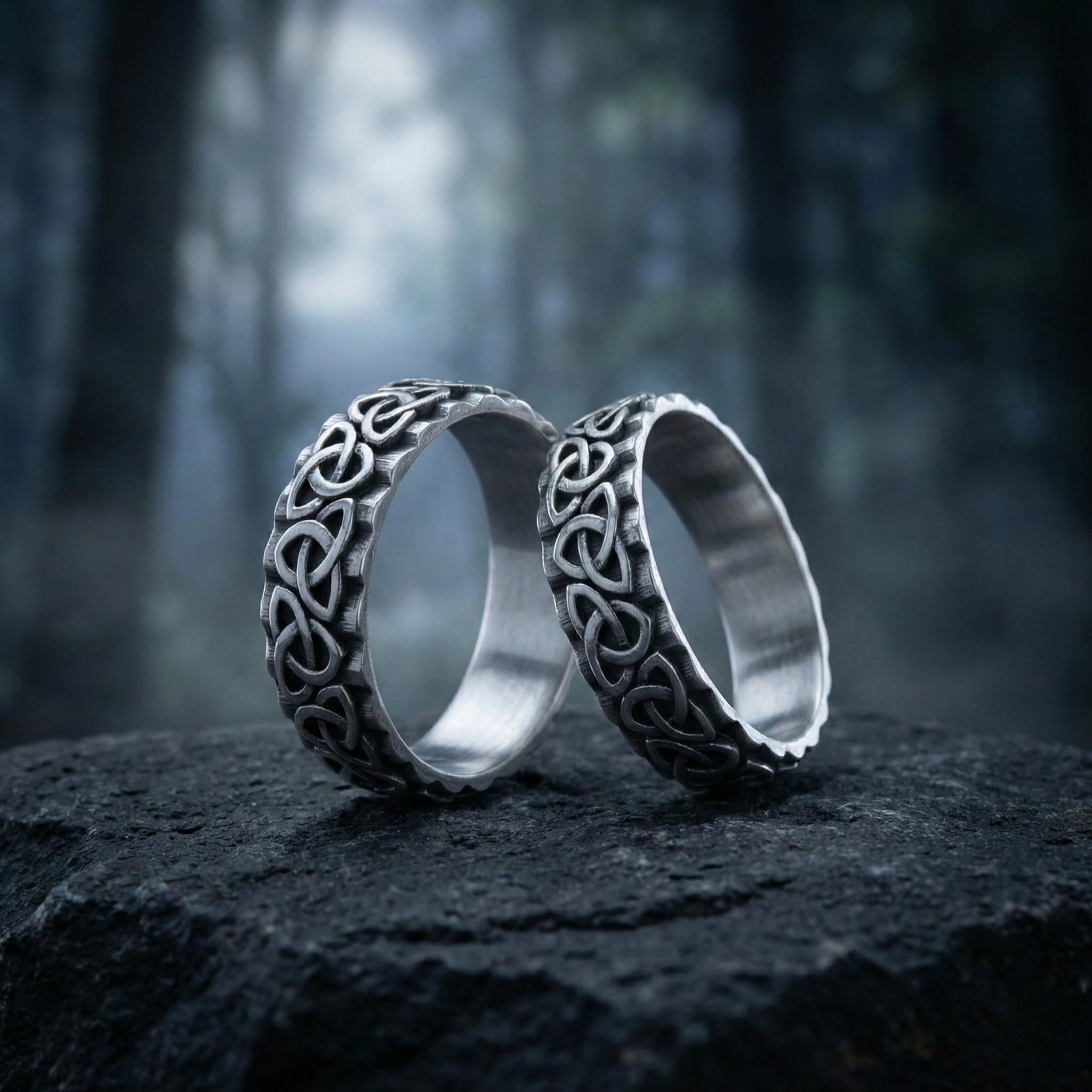 Two silver rings with intricate designs are placed on a dark surface, possibly a rock or a stone, against a blurred forest background.