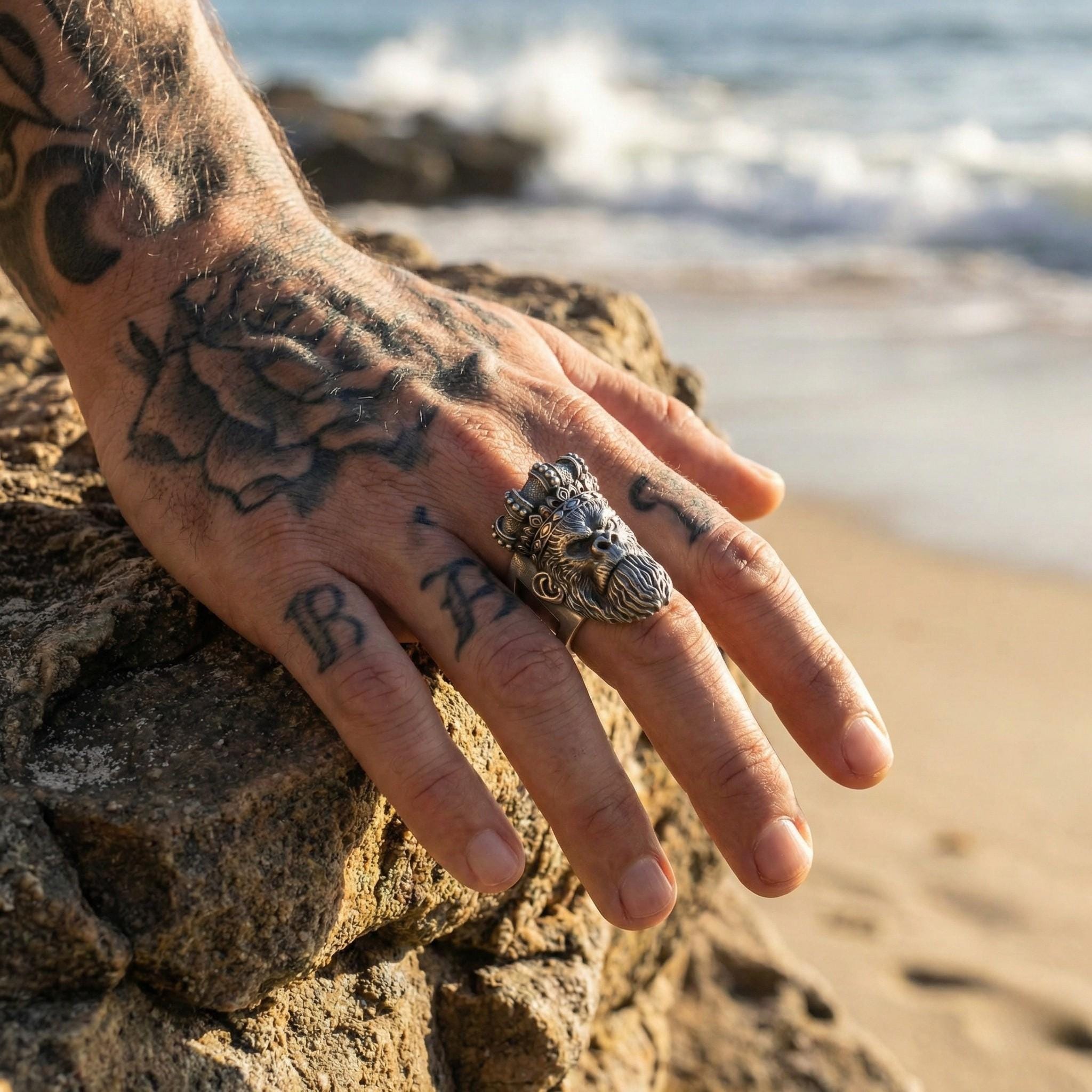A close-up of a person's hand with tattoos on the fingers and a large metal ring on the ring finger, with the ocean visible in the background.