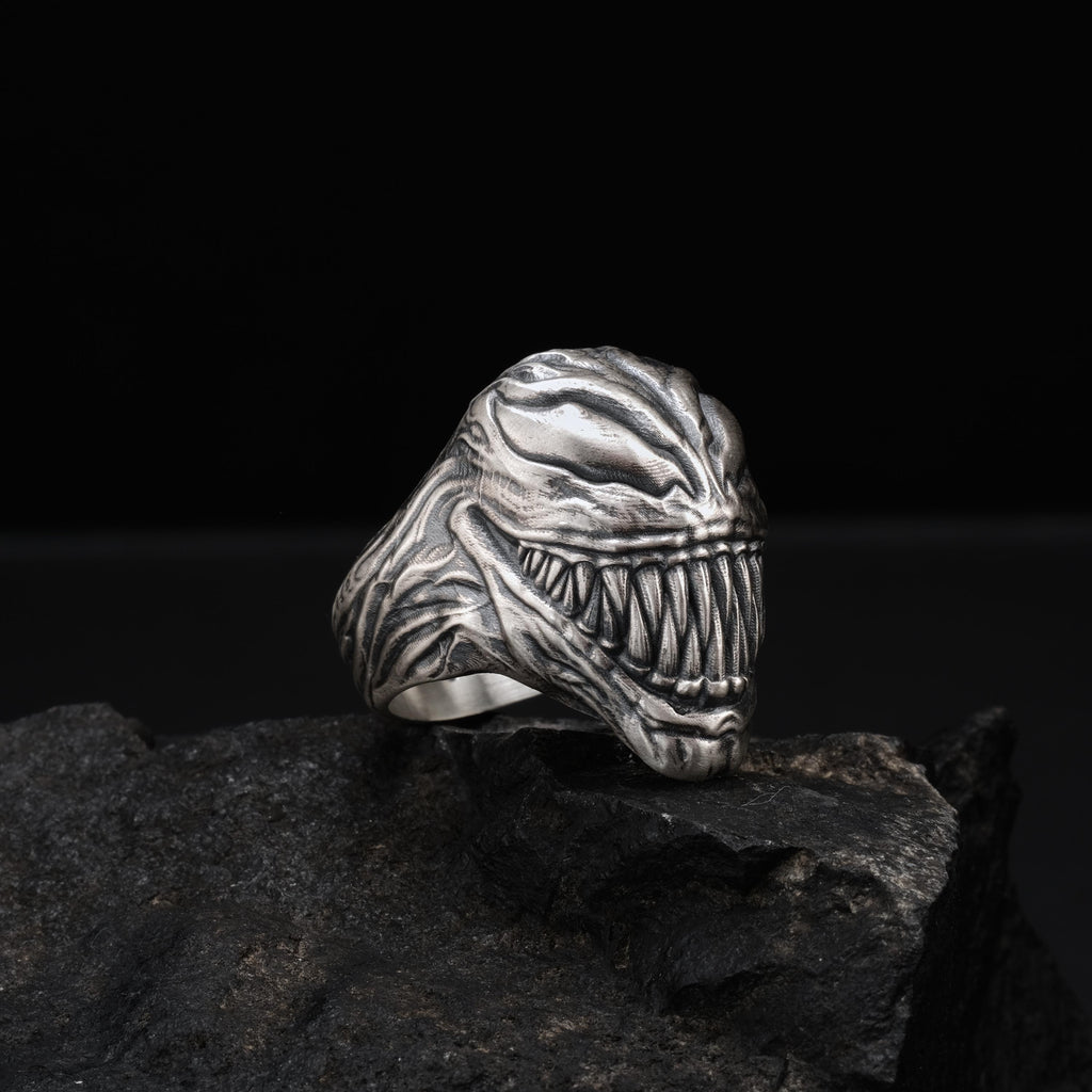 A silver ring with a menacing, monster-like design sits on a dark, rocky surface.