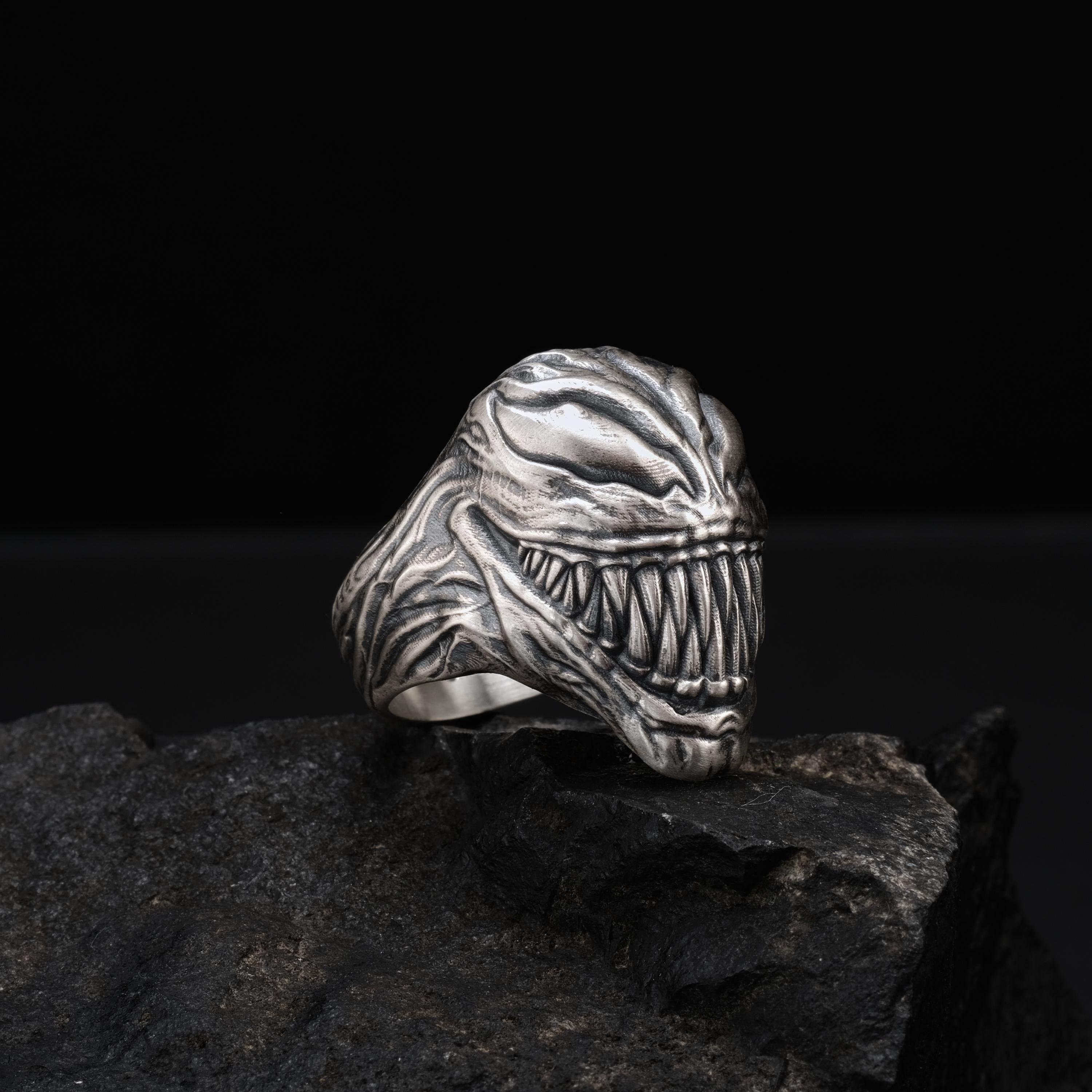 A silver ring with a menacing, monster-like design sits on a dark, rocky surface.