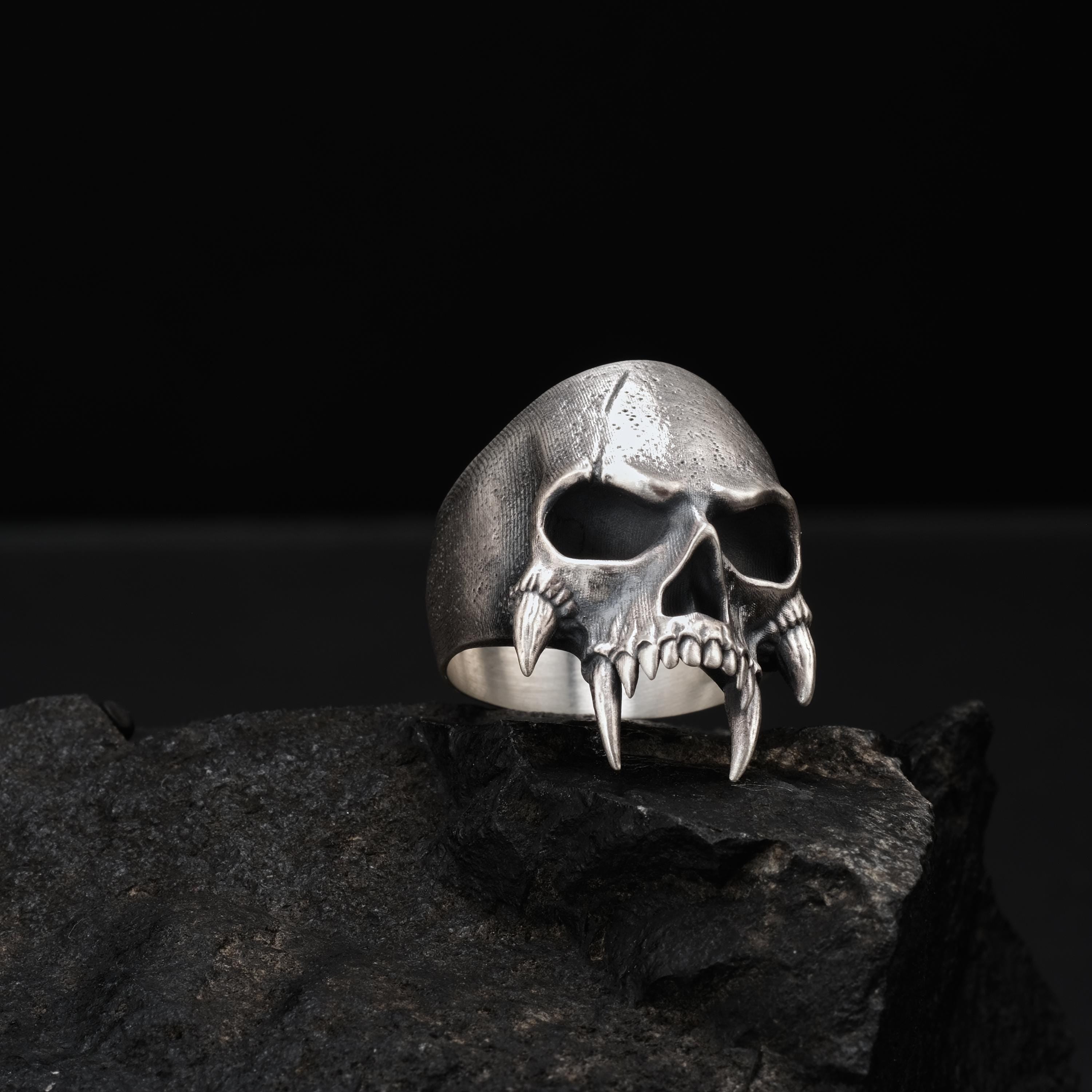 A silver skull ring with fangs is displayed on a rock against a dark background.