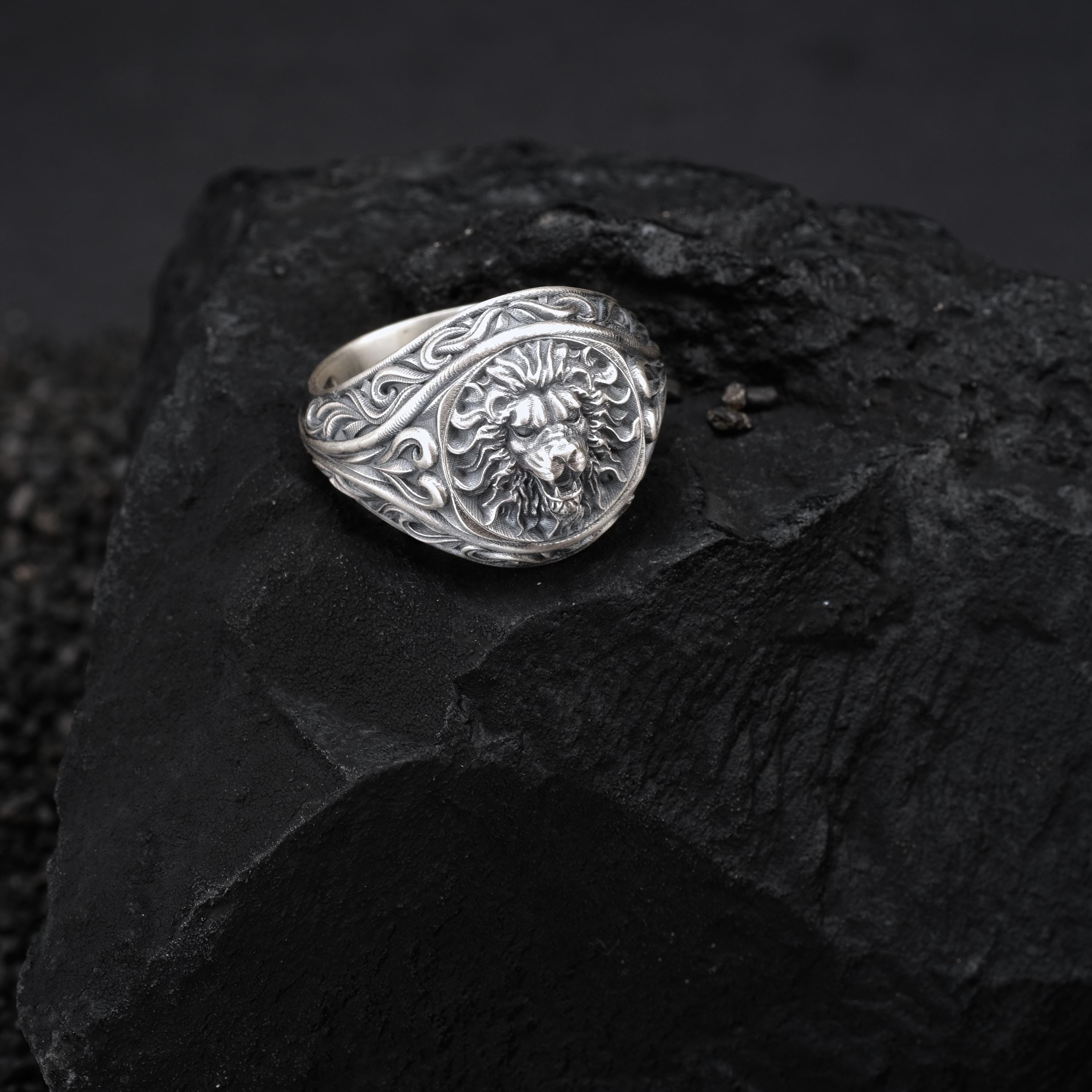 A silver ring with an intricate lion head design rests on a dark, rocky surface.