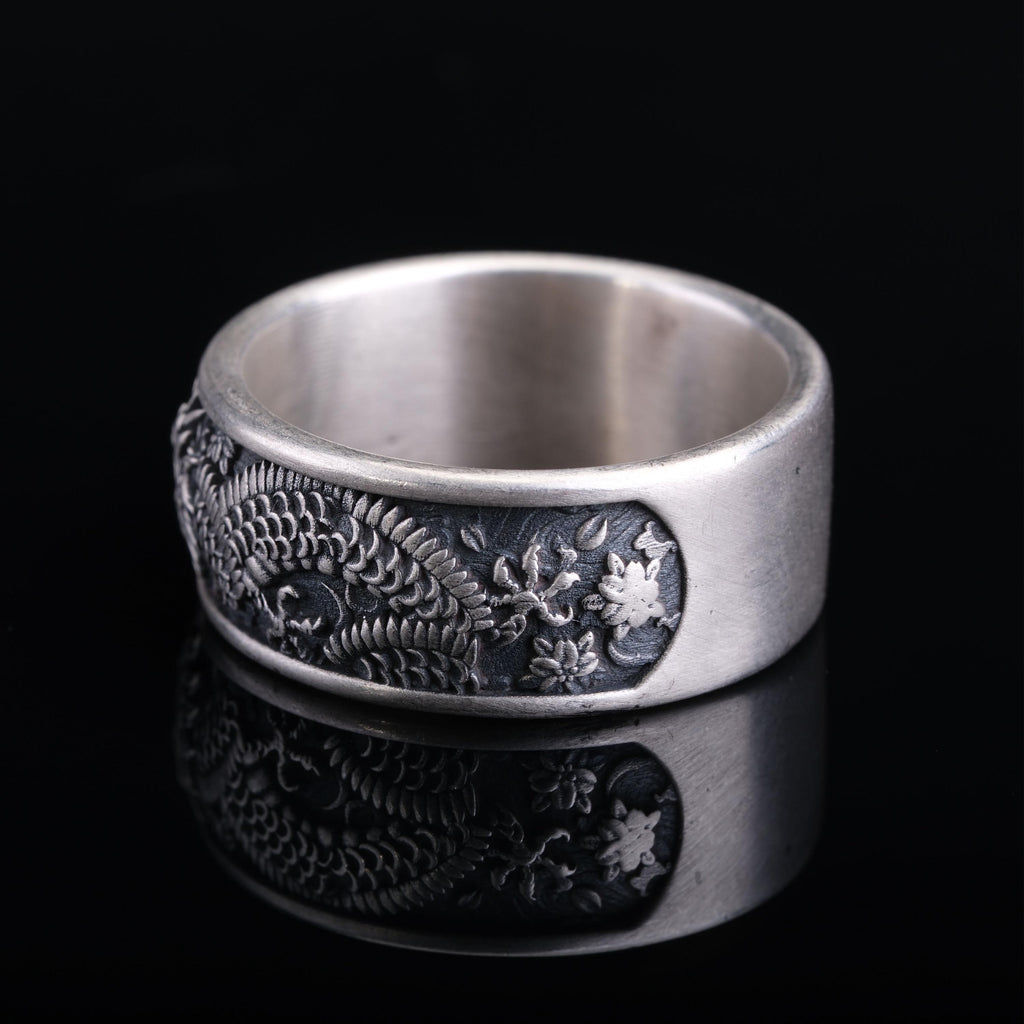 A silver ring with an intricate engraved design, featuring a dragon motif, is displayed against a black background.
