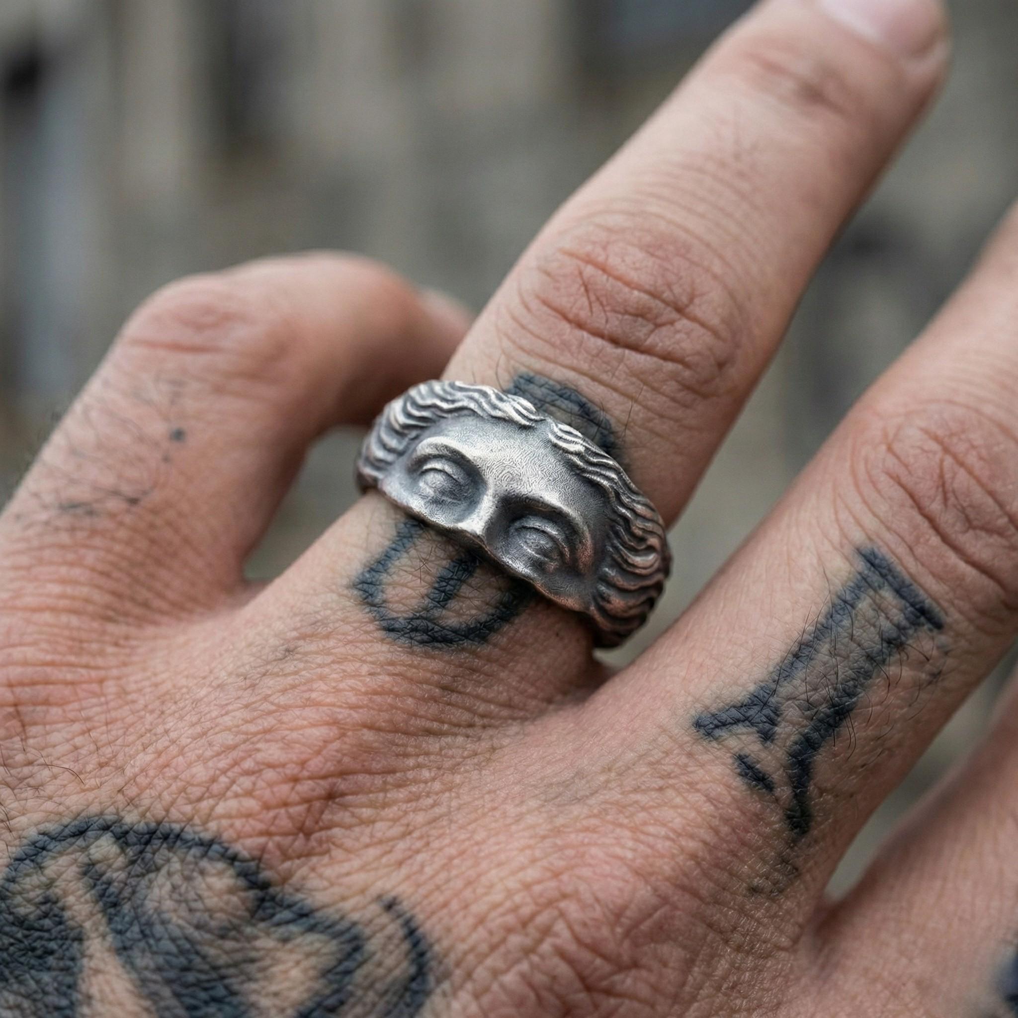 A close-up of a person's hand wearing a ring with a face design on it, and tattoos on the fingers.