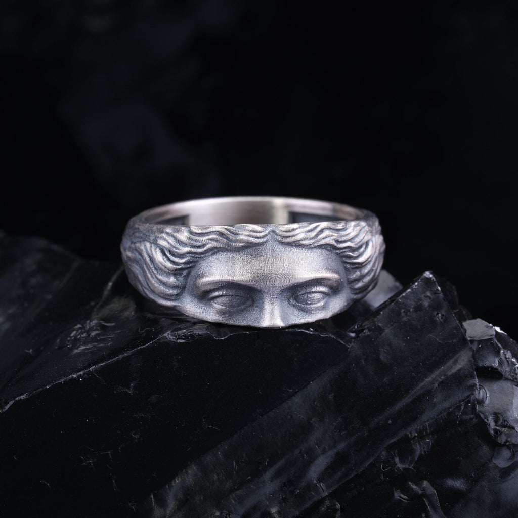 A silver ring with a face design on it, placed on a black surface.