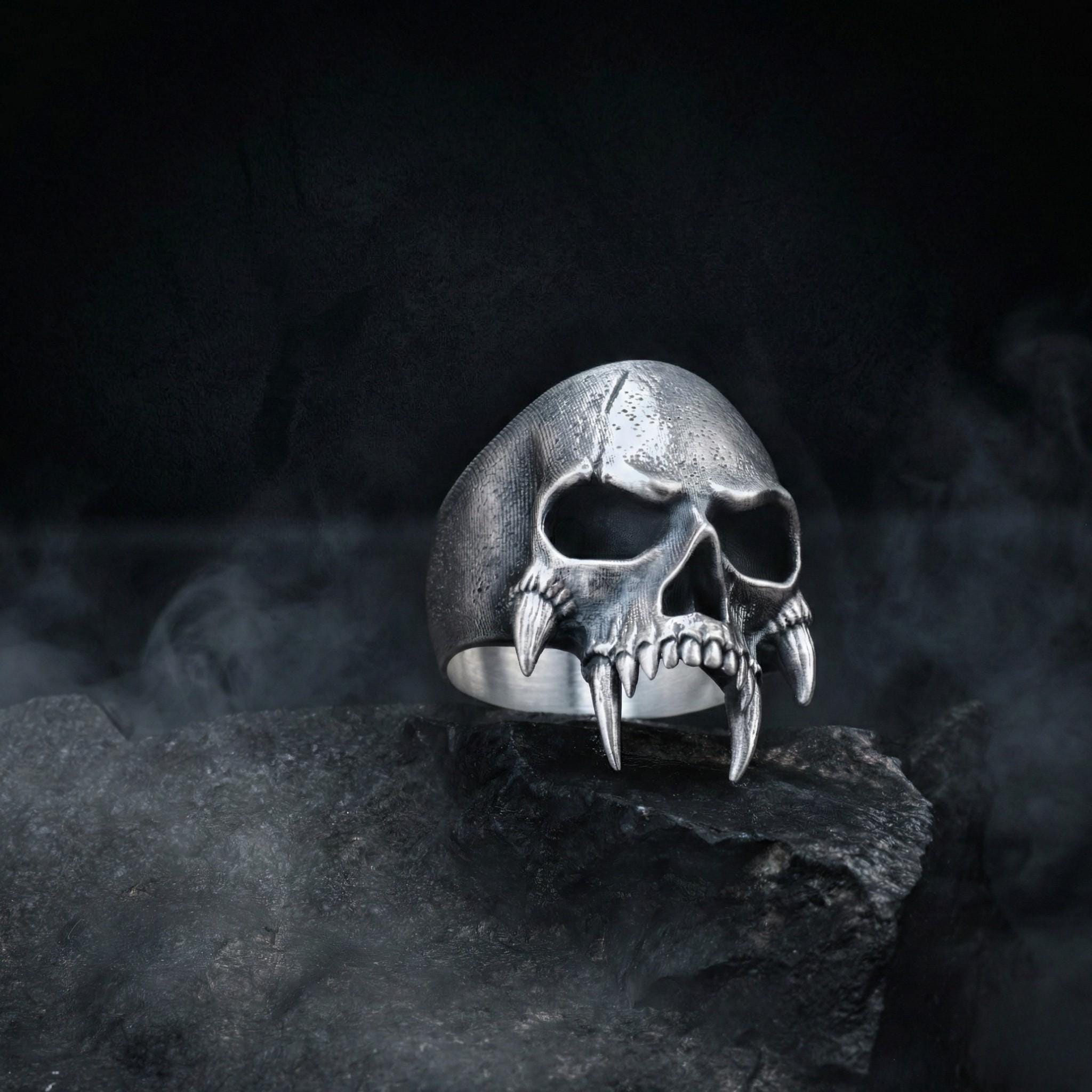 A silver skull ring with fangs is displayed against a dark, foggy background.