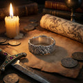a silver ring with intricate designs, a lit candle, a sword, coins, and a map with a dragon on it, all arranged on a wooden surface.
