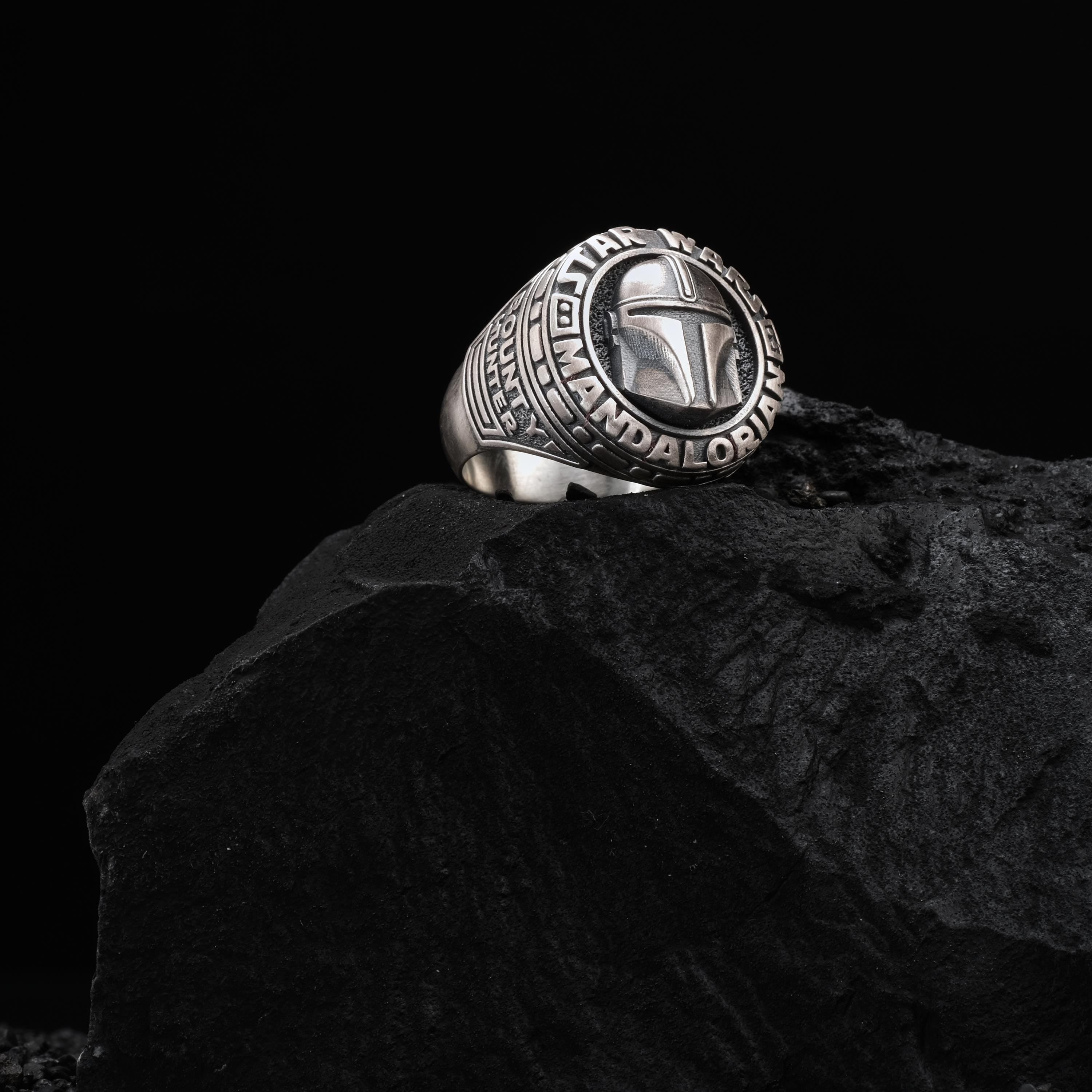 A silver ring with a Star Wars-themed design sits atop a dark, rocky surface.