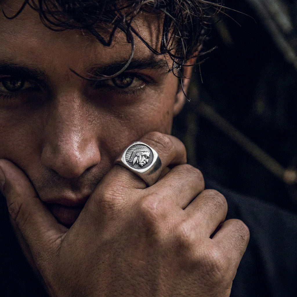 A close-up of a man's face, with his eyes looking directly at the camera. He is wearing a silver ring with an image of a Native American head on it.