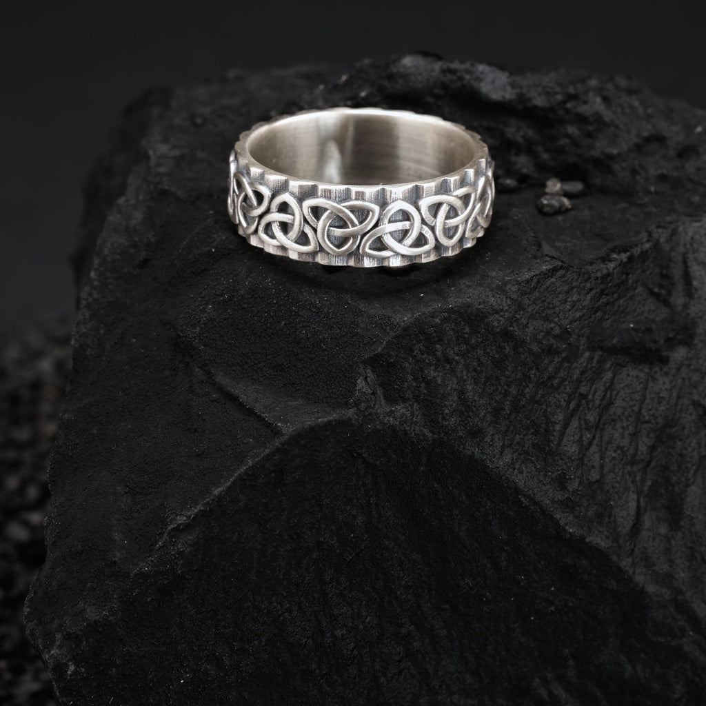 A silver ring with an intricate Celtic knot design sits on a dark, rocky surface.