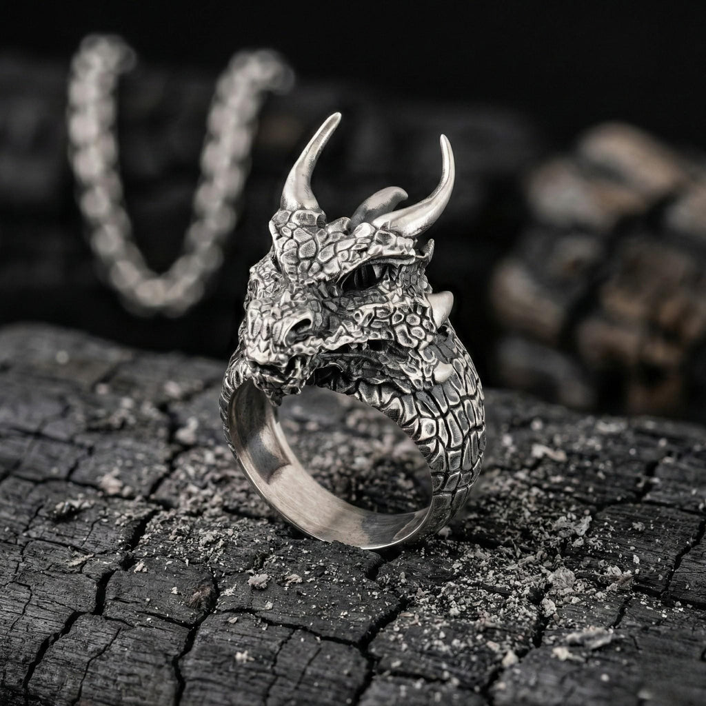 A silver ring with a dragon head design sits on a dark, textured surface, with a chain visible in the background.
