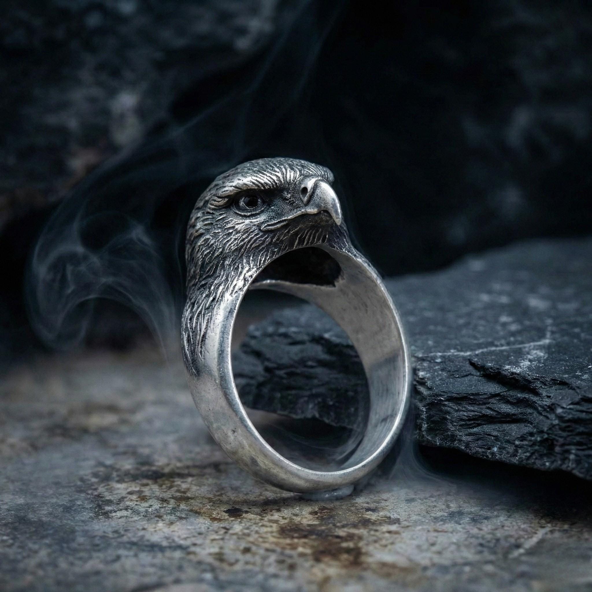 A silver ring with an eagle head design is shown against a dark background, with smoke or fog emanating from it.