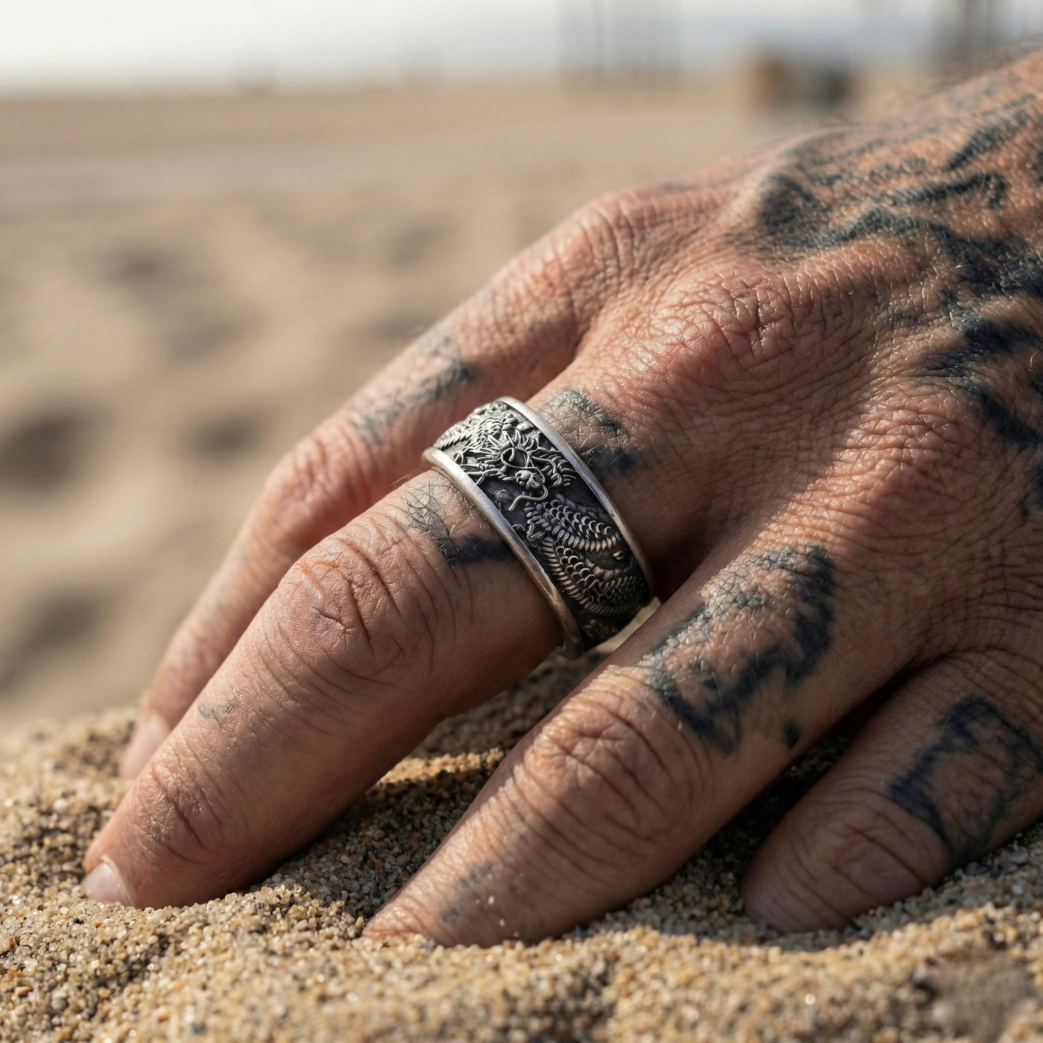 A close-up of a person's hand wearing a silver ring with a dragon design, resting on a sandy beach.