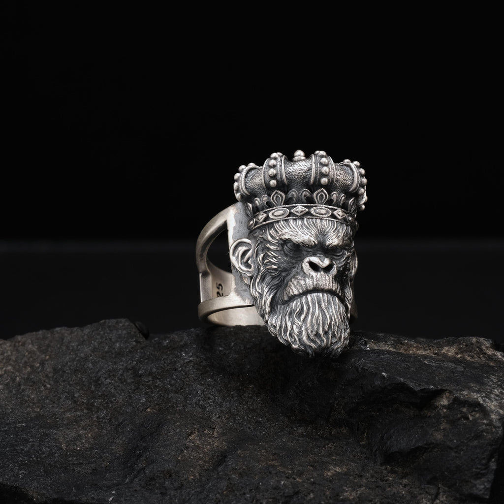 A silver ring featuring a detailed, carved monkey head wearing a crown, placed on a dark surface.