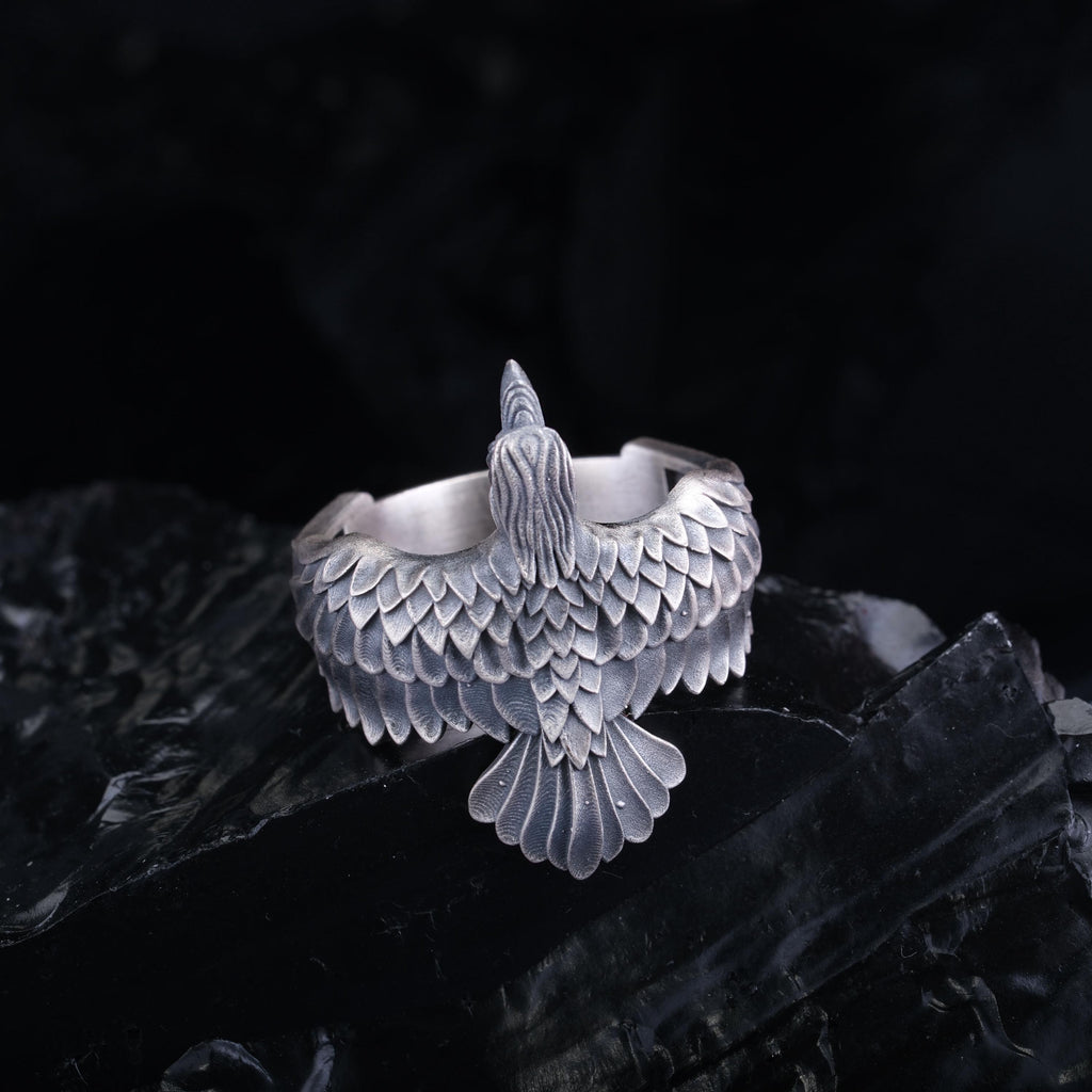 A silver ring with a detailed bird design, featuring a large beak and intricate feather patterns, is displayed against a dark background.