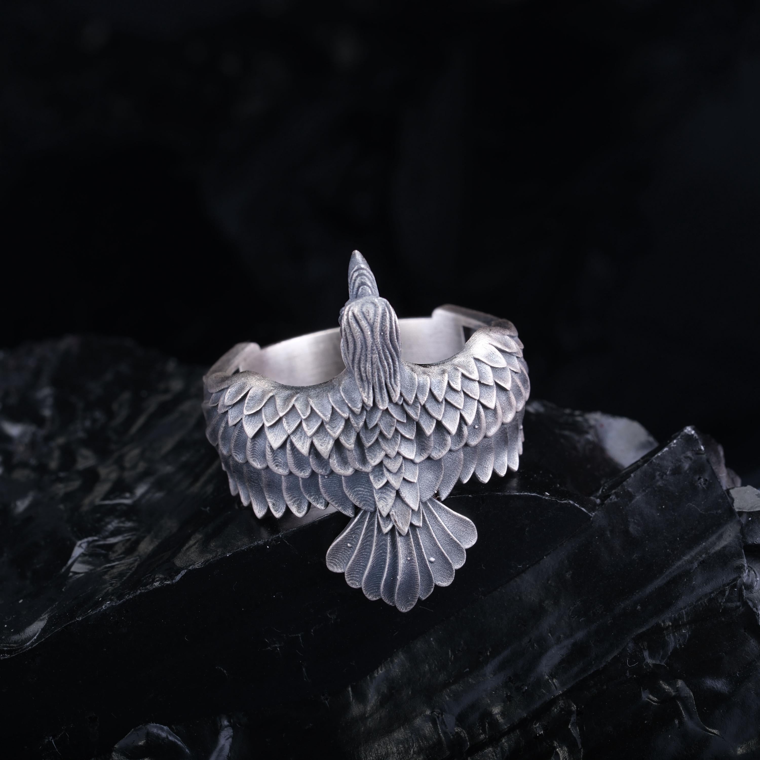 A silver ring with a detailed bird design, featuring a large beak and intricate feather patterns, is displayed against a dark background.