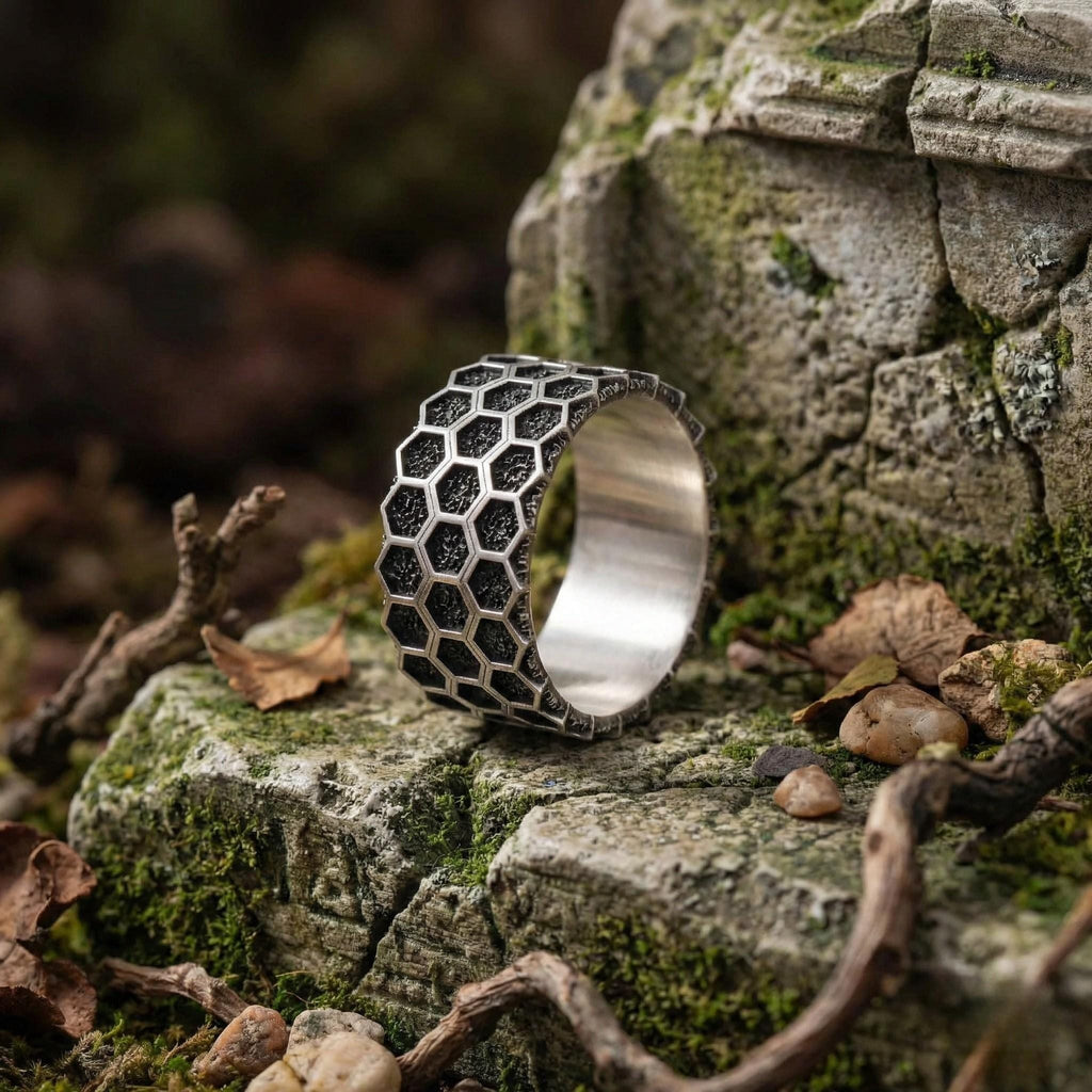 A silver ring with a honeycomb pattern sits on a mossy rock in a forest setting.