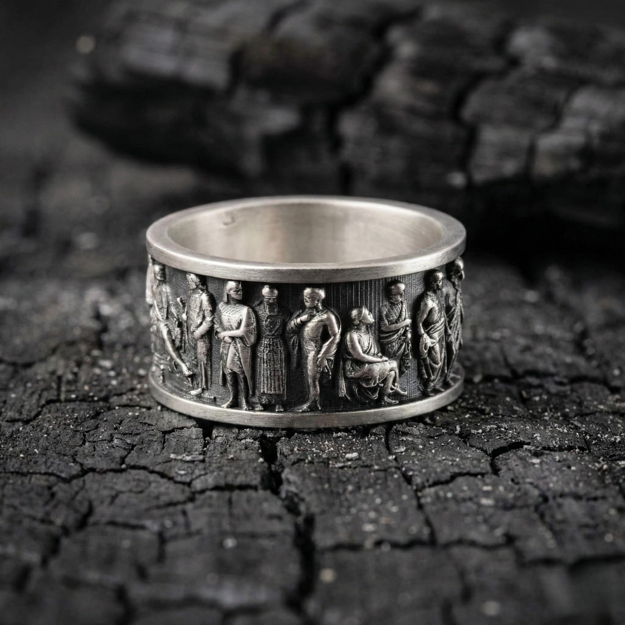 A silver ring with a detailed engraved design featuring various figures, including men and women, on a dark, cracked surface.