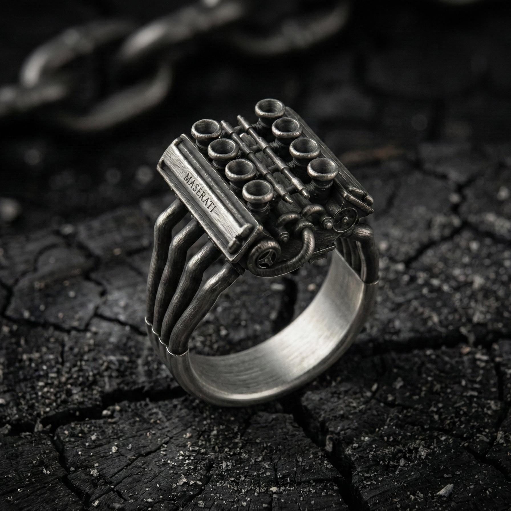 A silver ring with an intricate design, resembling an engine or machinery, is displayed on a dark, cracked surface.