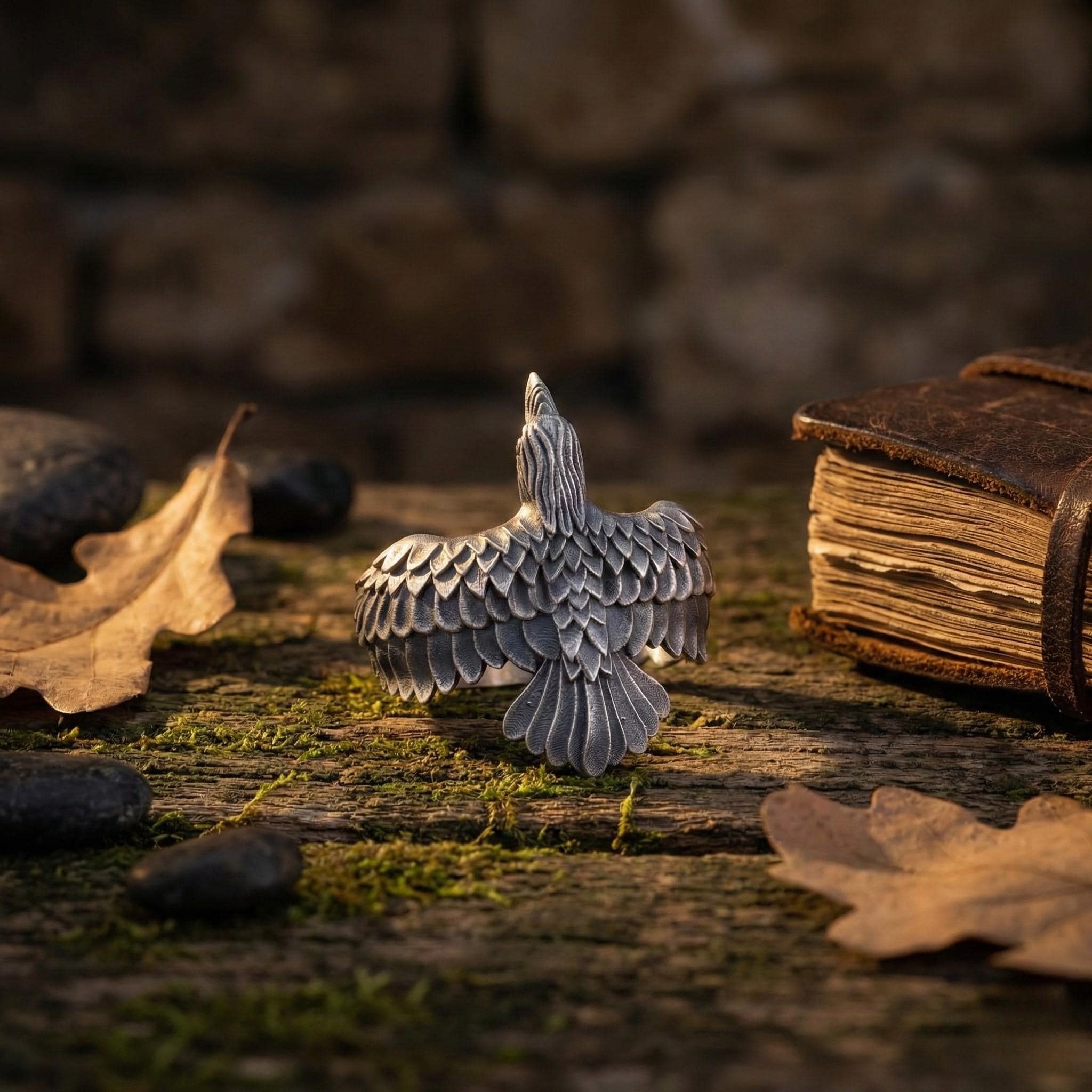 a silver bird sculpture with outstretched wings, surrounded by fallen leaves and rocks on a mossy surface.