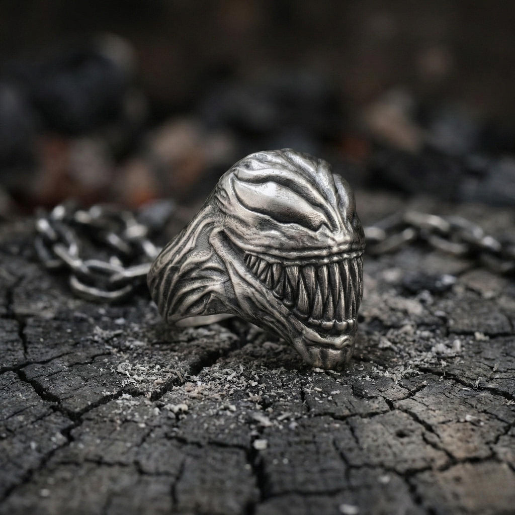 A close-up image of a silver ring with a menacing, monster-like design, resting on a cracked, black surface.
