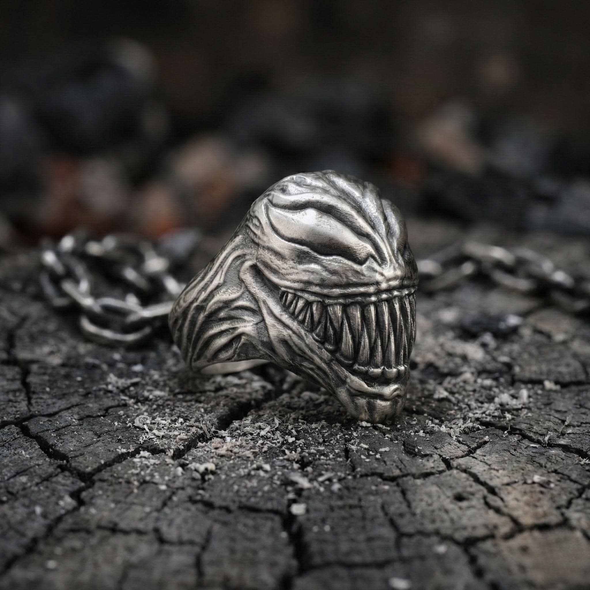 A close-up image of a silver ring with a menacing, monster-like design, resting on a cracked, black surface.