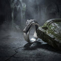 A silver ring with a dragon head design is resting on a moss-covered stone surface, with a dark, foggy background.