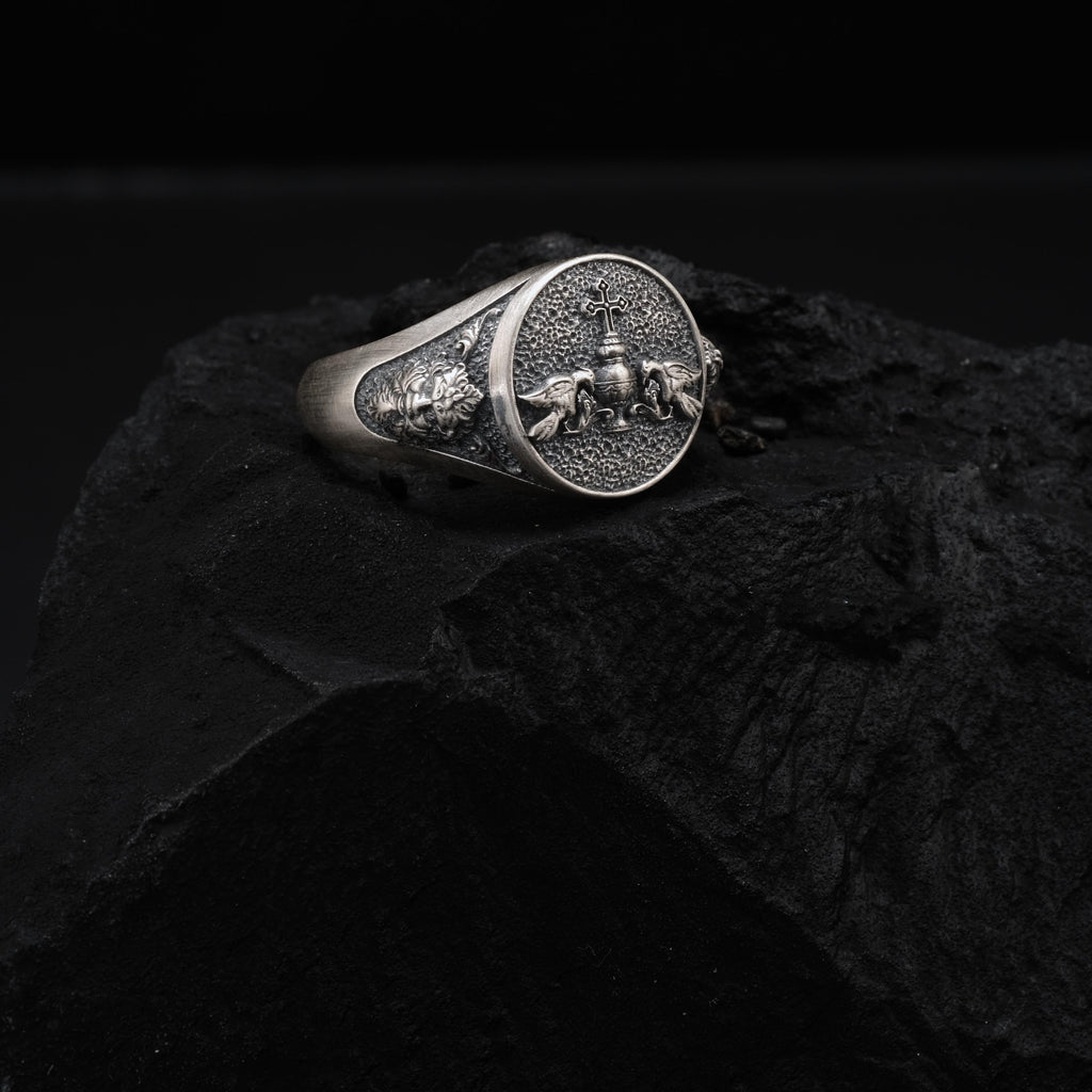 A silver ring with an embossed design sits on a dark surface, possibly a rock or a piece of stone.