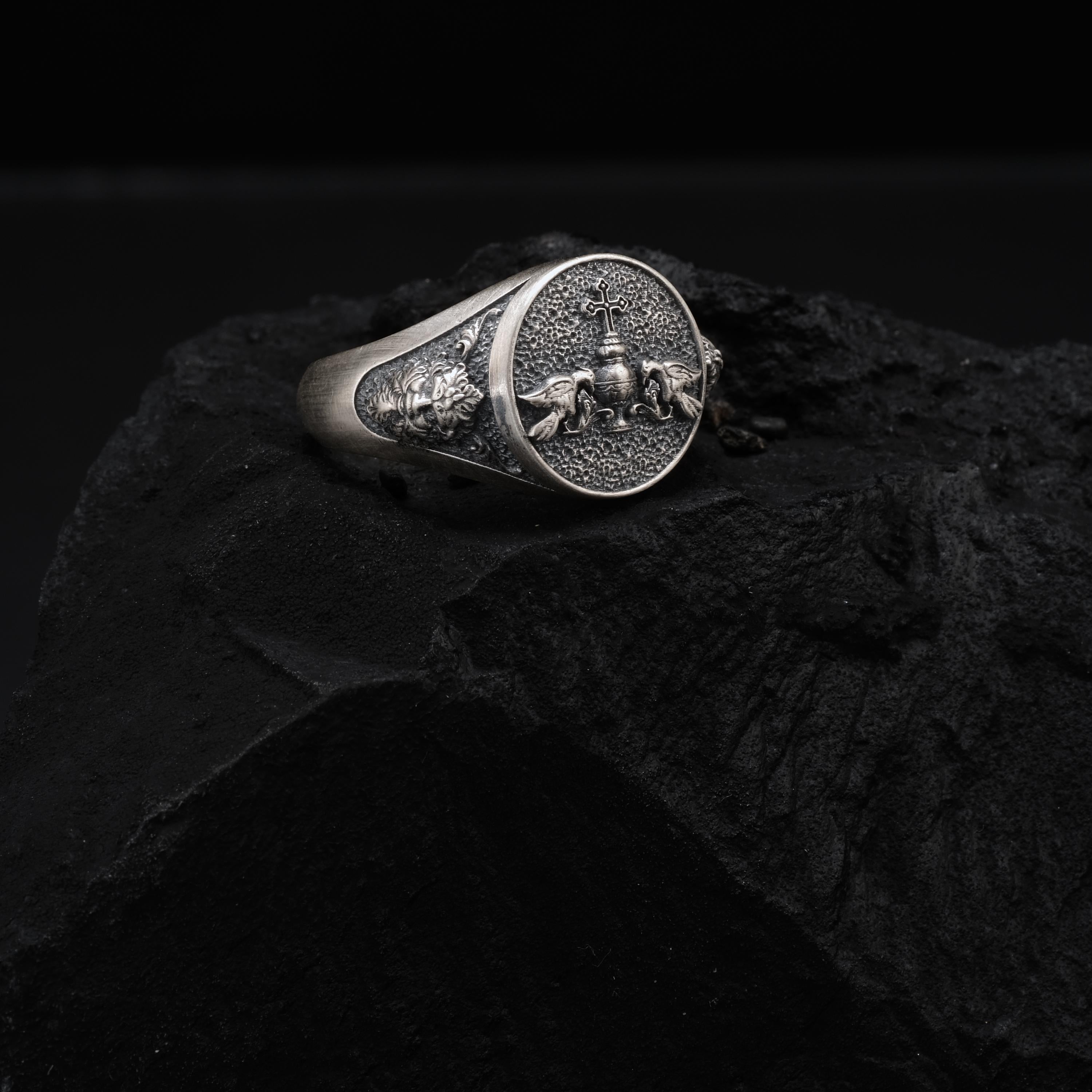 A silver ring with an embossed design sits on a dark surface, possibly a rock or a piece of stone.