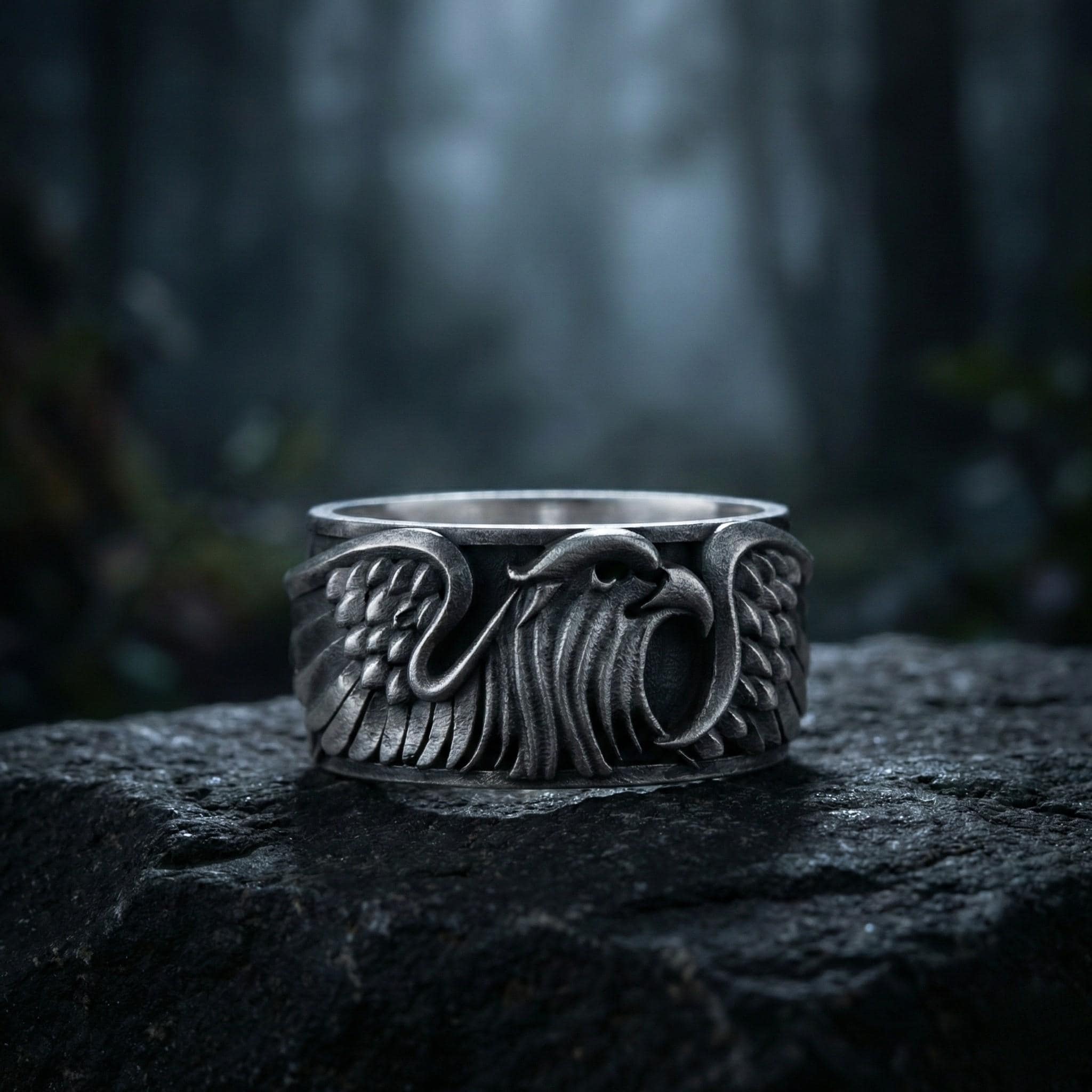 A silver ring with an intricate design featuring a winged eagle or bird-like figure, set against a dark, blurred background.