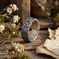 A silver ring with a floral design sits on a wooden surface, surrounded by dried flowers and a piece of paper.