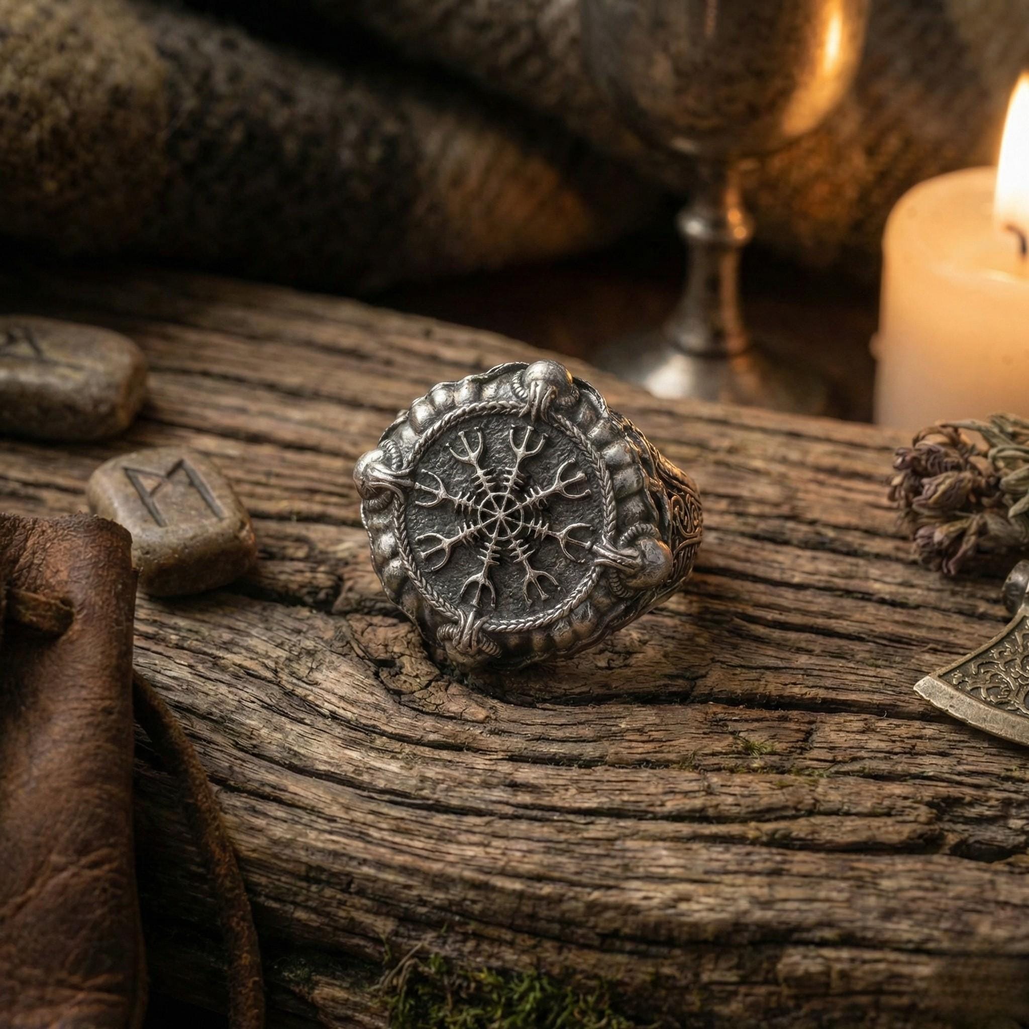 a silver ring with a detailed design, placed on a wooden surface. The ring is surrounded by various objects, including a candle, a leather item, and what appears to be a piece of jewelry.