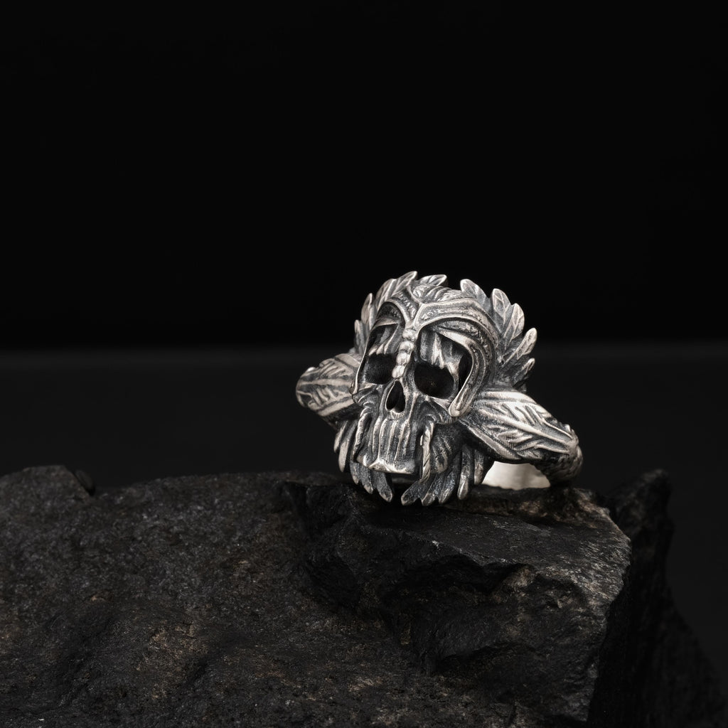 A silver skull ring with intricate leaf designs sits on a dark rock.