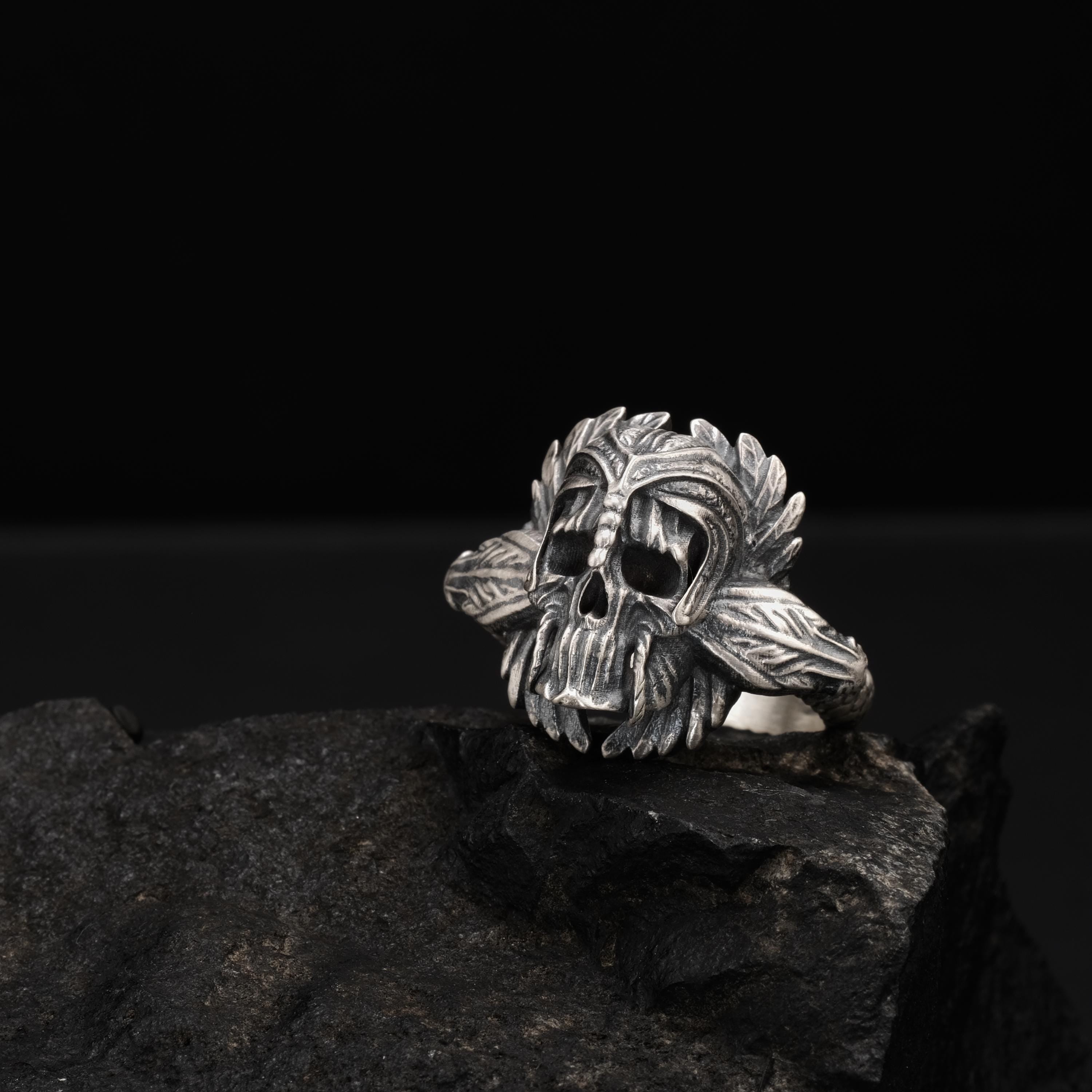 A silver skull ring with intricate leaf designs sits on a dark rock.