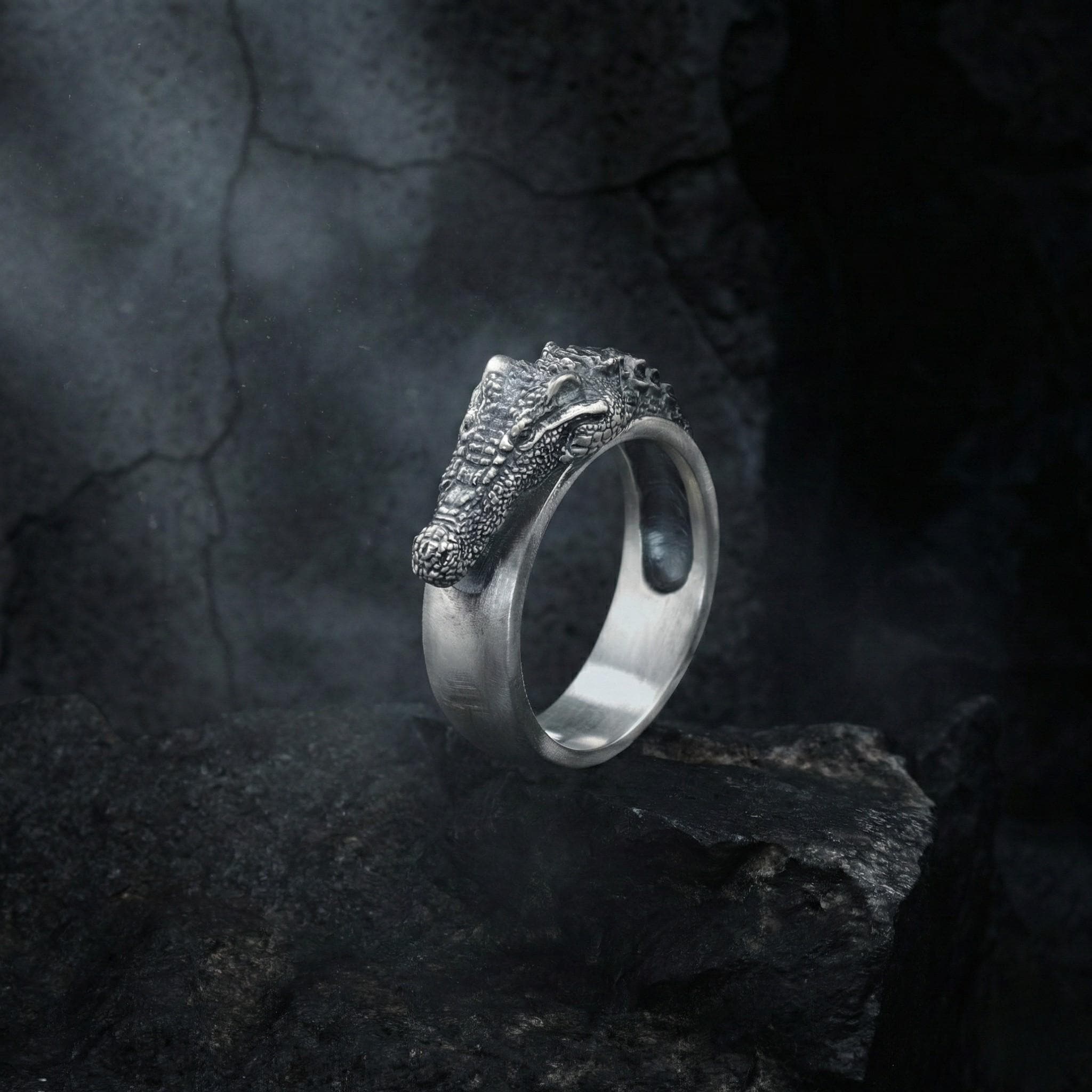 A silver ring with a crocodile head design is displayed against a dark, rocky background.