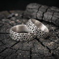 Two silver rings with intricate Celtic knot designs are resting on a dark, textured surface.