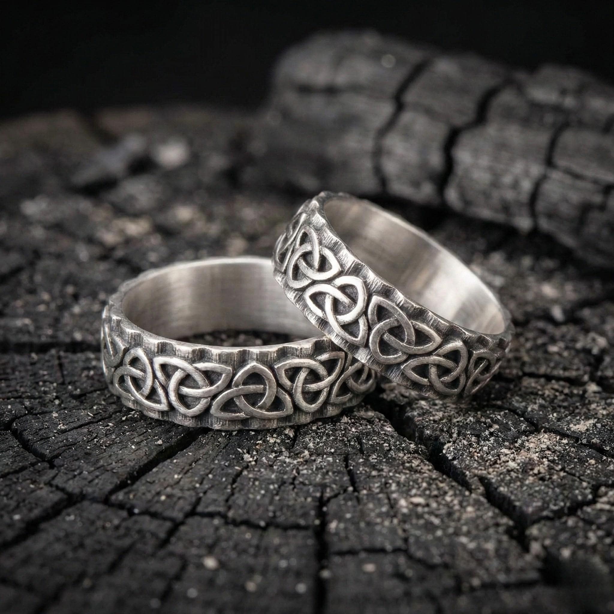 Two silver rings with intricate Celtic knot designs are resting on a dark, textured surface.