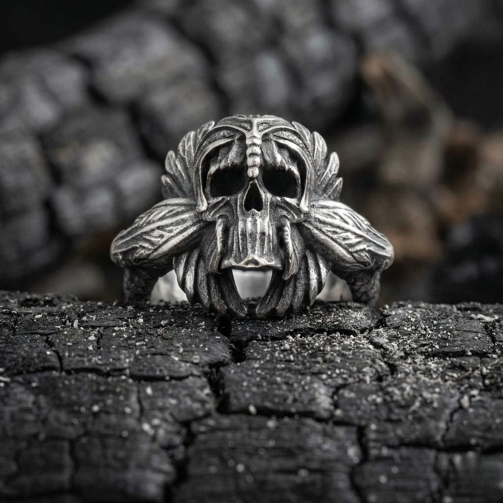 A silver skull ring with intricate designs, resting on a black, cracked surface.