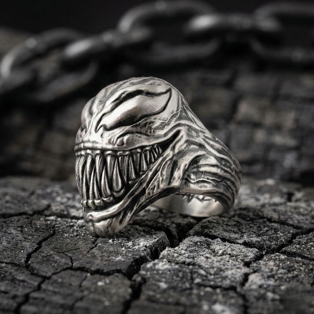 A close-up of a silver ring with a menacing, monster-like design, featuring sharp teeth and an ominous expression, resting on a cracked, weathered surface.