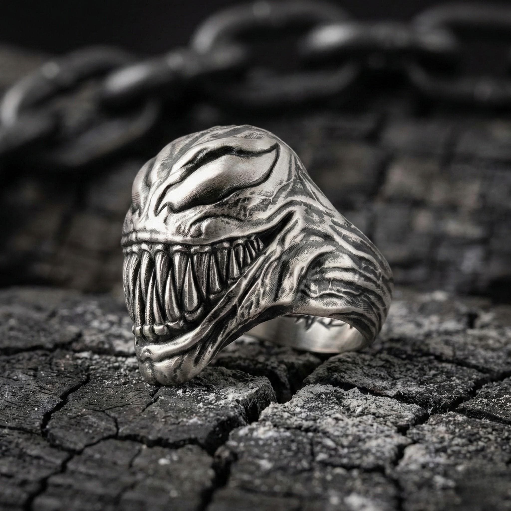 A close-up of a silver ring with a menacing, monster-like design, featuring sharp teeth and an ominous expression, resting on a cracked, weathered surface.