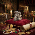 a collection of ornate and valuable items, including a crown, a large metal object resembling a monkey's head, a rolled scroll, and various coins and jewels, all arranged on a red velvet surface.