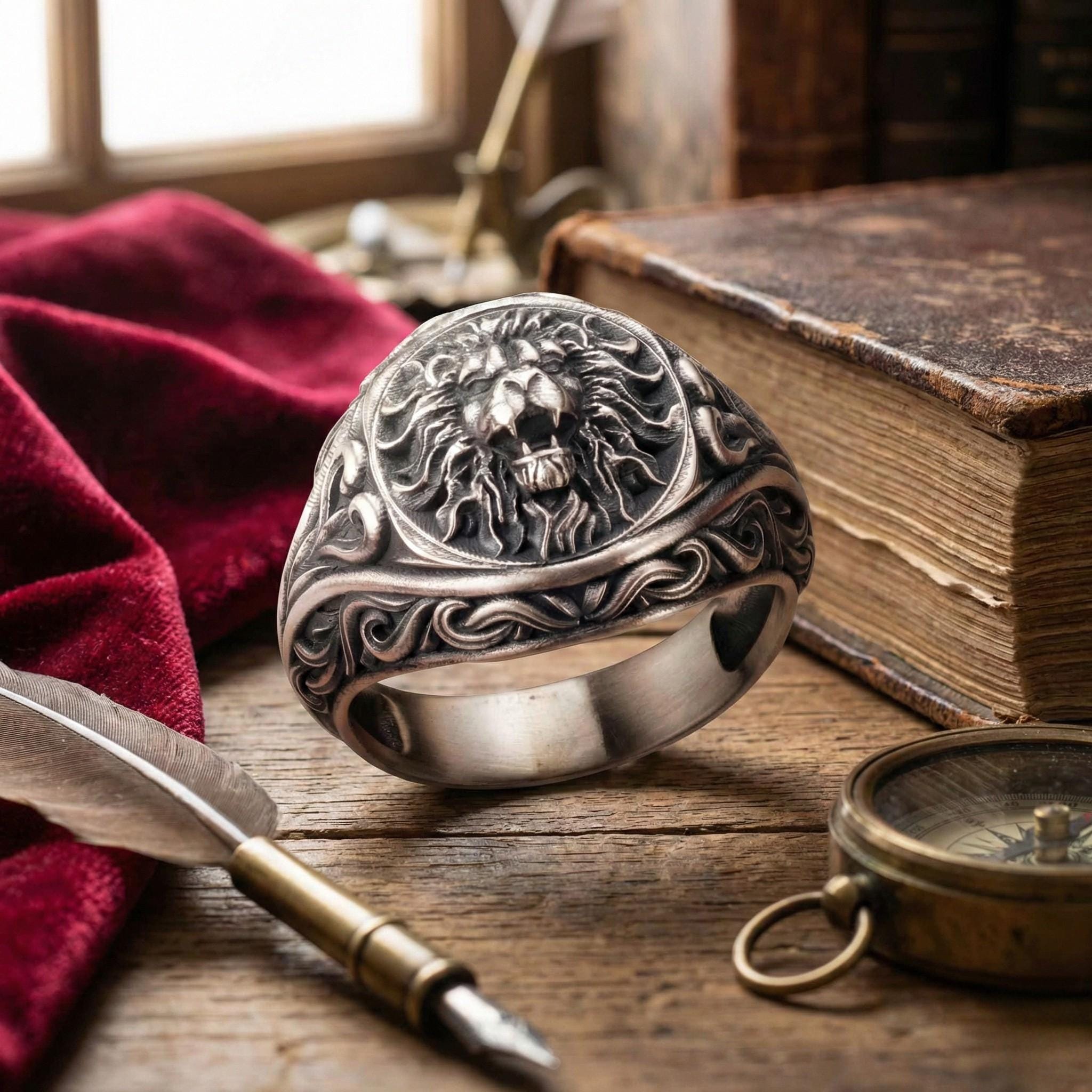 a silver ring with an intricate lion design, a feather quill pen, a compass, and a book on a wooden surface.