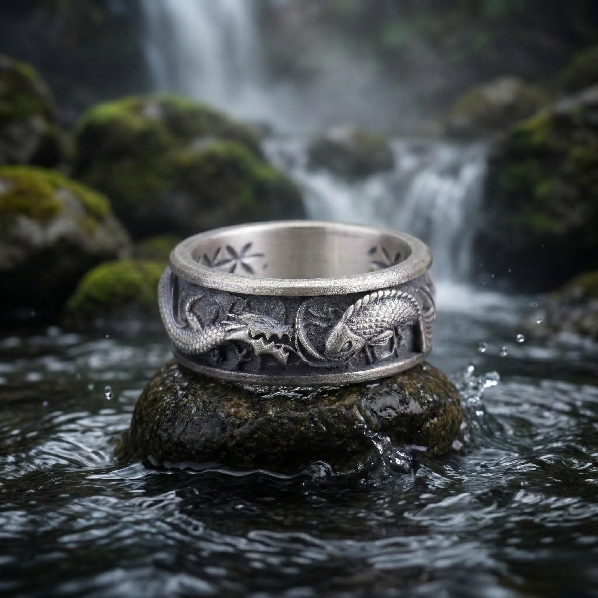 A silver ring with a dragon design sits on a rock in a stream, surrounded by mossy rocks and a waterfall in the background.