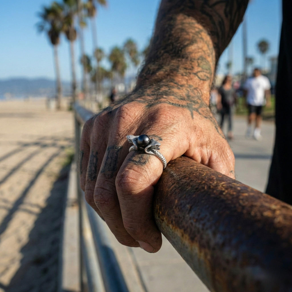 A tattooed hand wearing a ring is holding onto a metal railing, with palm trees and a beach visible in the background.