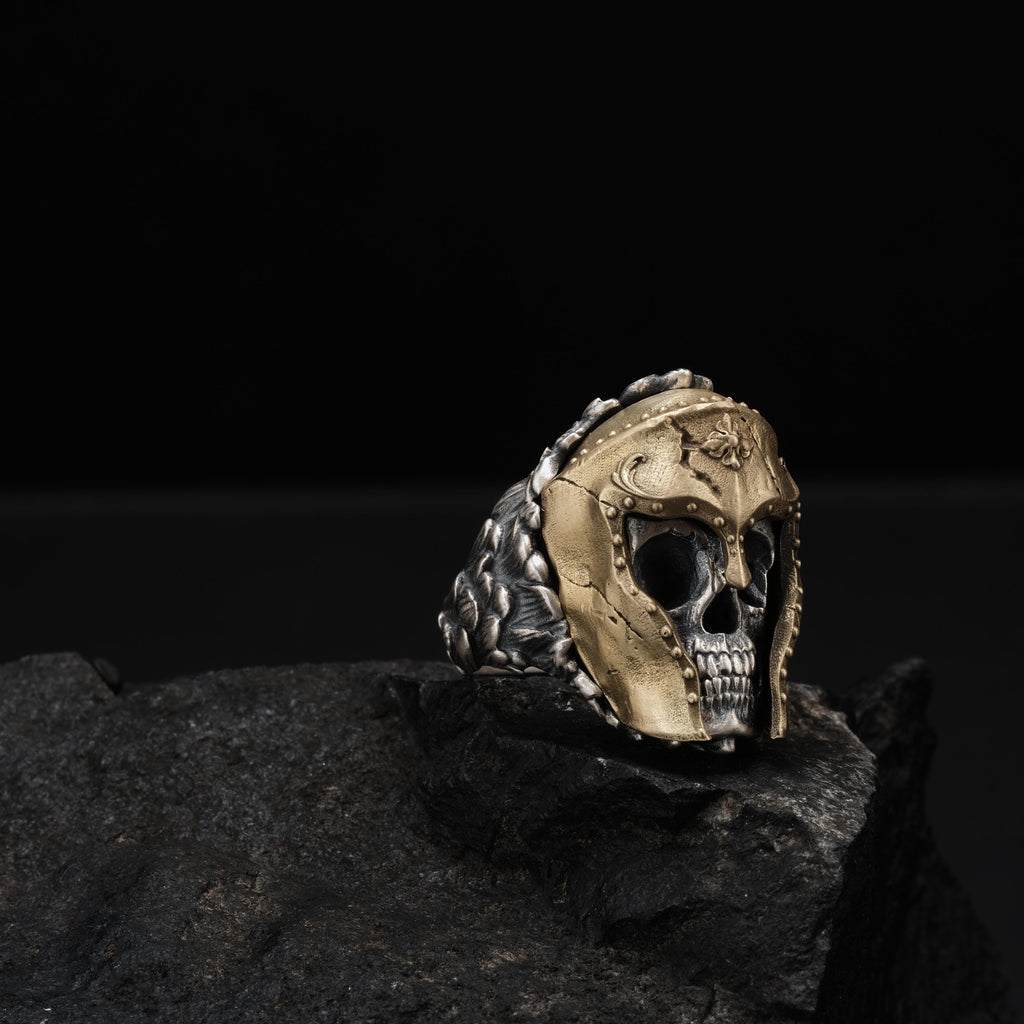 A gold-colored ring with a skull-like design sits atop a dark rock, creating a striking contrast.