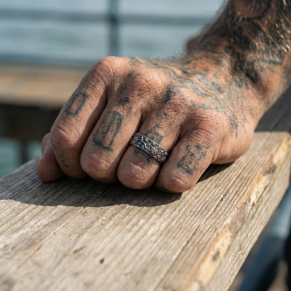 A close-up of a tattooed hand resting on a wooden surface, with a ring visible on one of the fingers.
